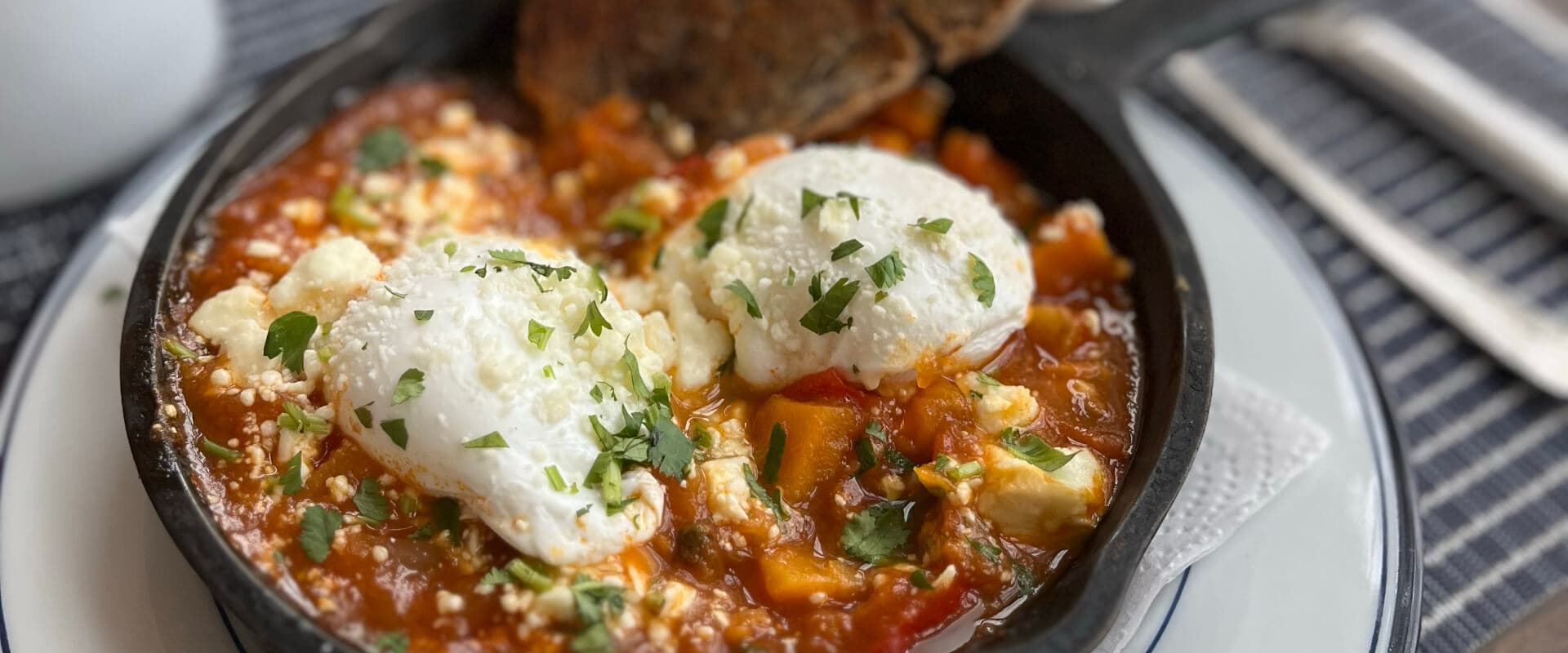 A cast-iron skillet filled with shakshuka topped with poached eggs and herbs, accompanied by a slice of bread.