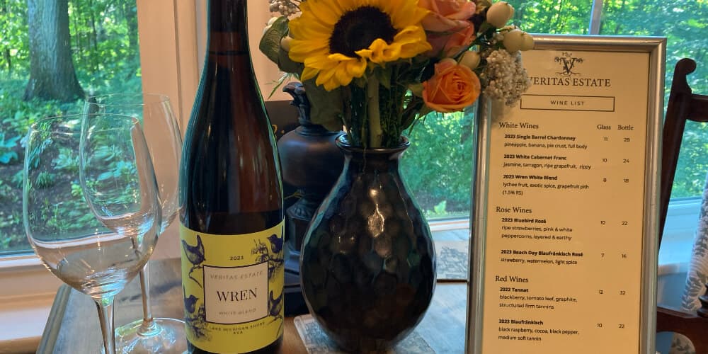 A bottle of Wren wine, a vase of sunflowers and other flowers, and a wine list are arranged on a table with a green view in the background.