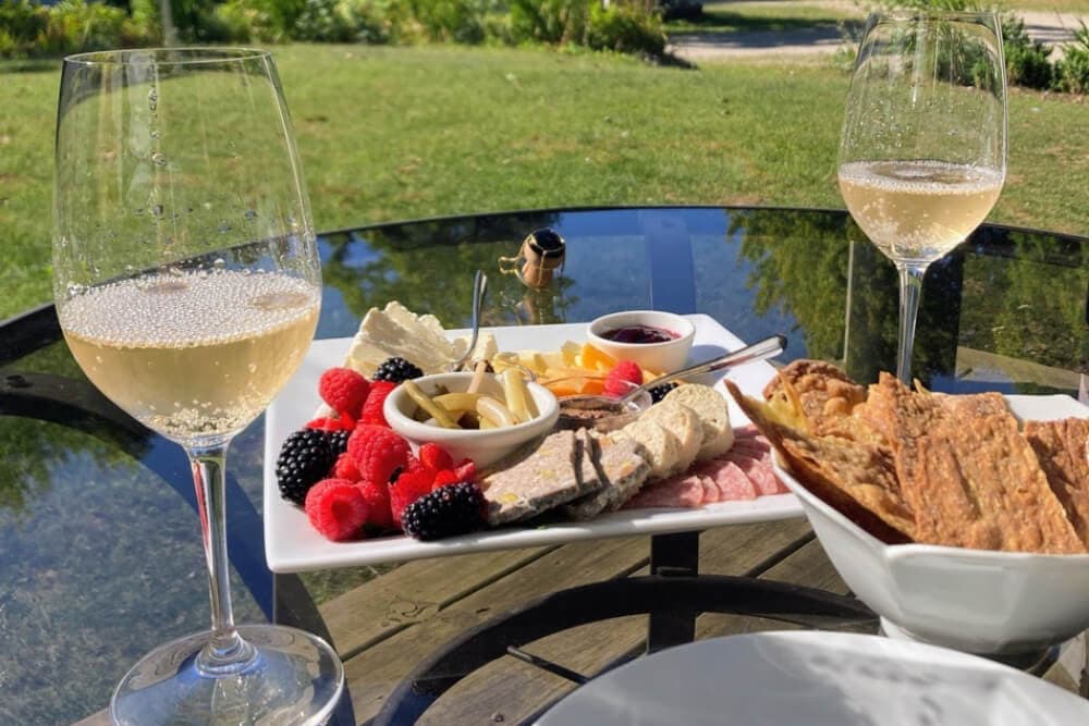 A table is set outdoors with two glasses of sparkling wine and a platter of fruits, cheeses, and meats.
