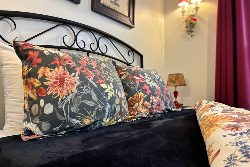 A bed with floral-patterned pillows and a dark blanket, accented by a nearby lamp and curtains.