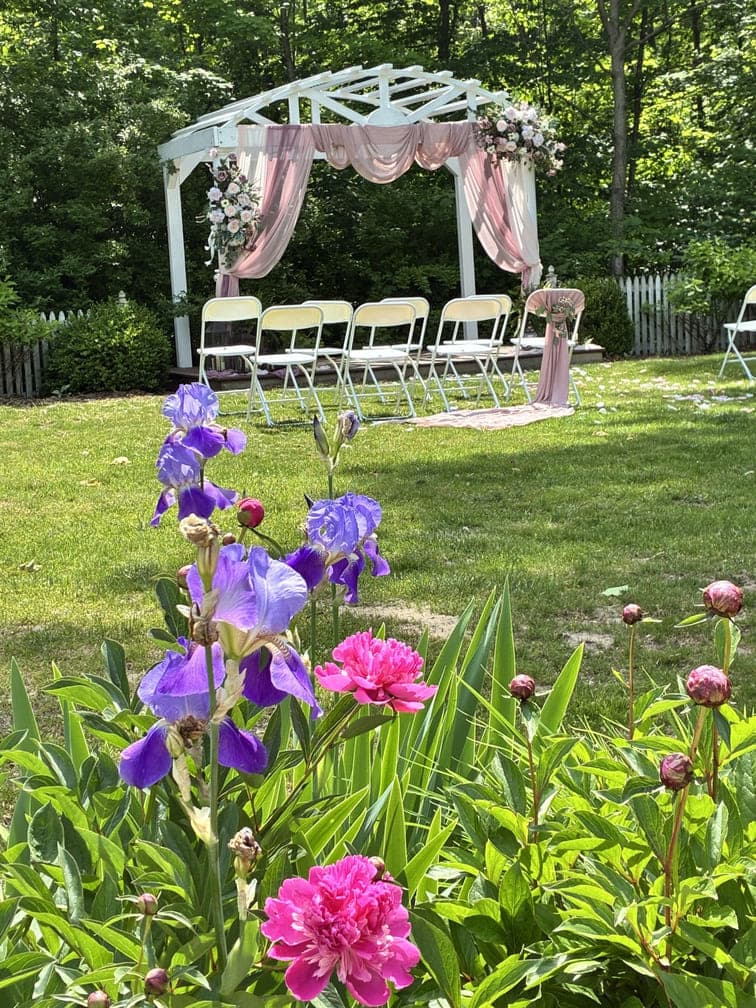 A floral garden setting with purple and pink flowers in the foreground and a decorated white arch with seating in the background, ideal for a wedding.