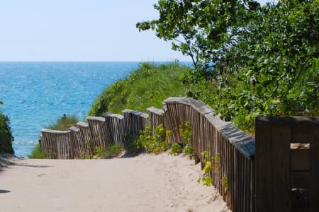 A sandy path leads to a tranquil beach with green vegetation and blue water.