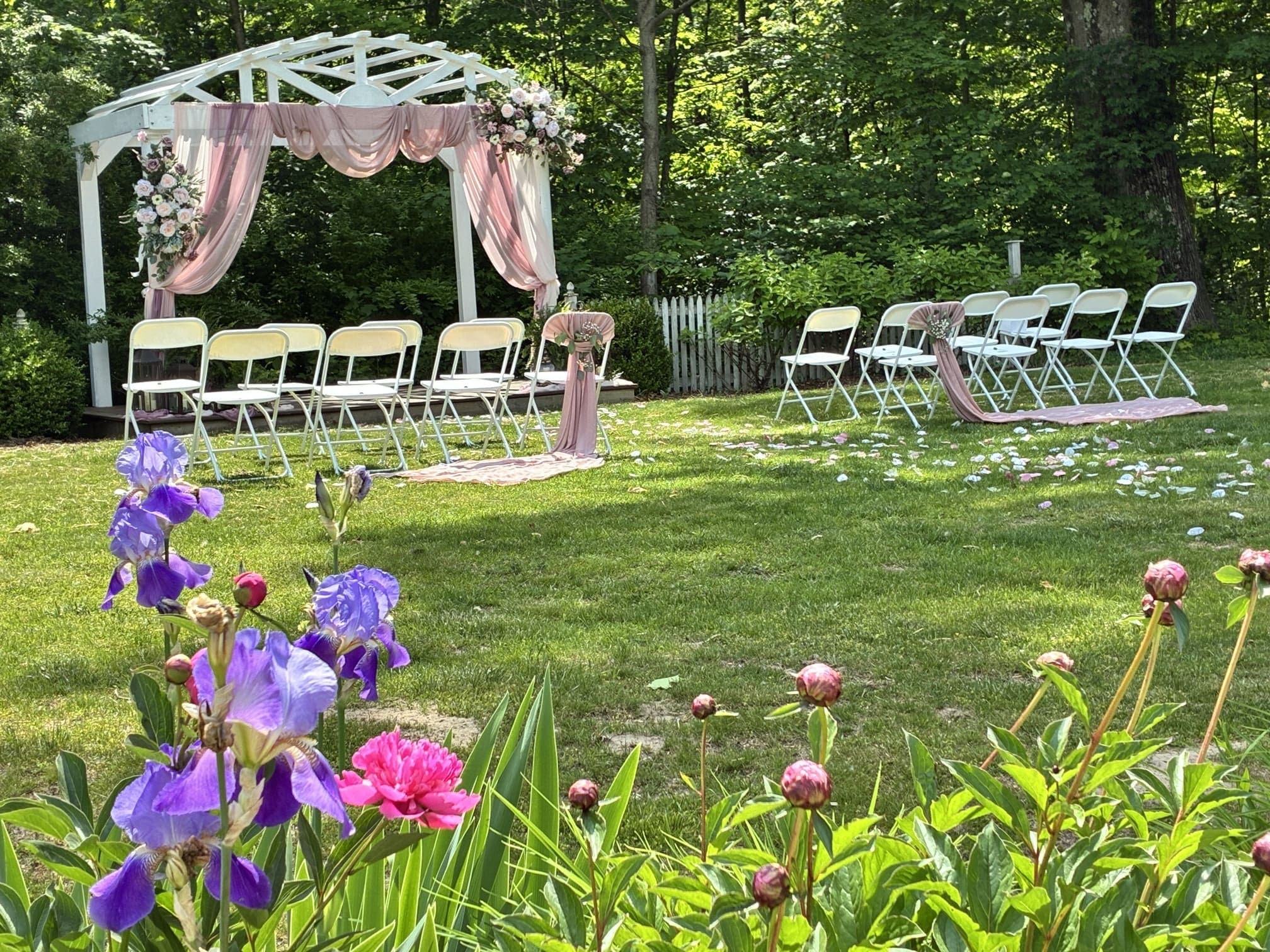 A serene outdoor wedding setup featuring a decorated arch and white chairs on a grassy lawn framed by colorful flowers.
