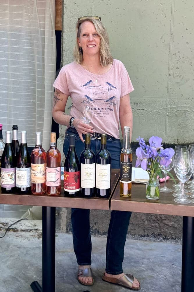 A woman stands behind a table displaying various wine bottles and flowers.