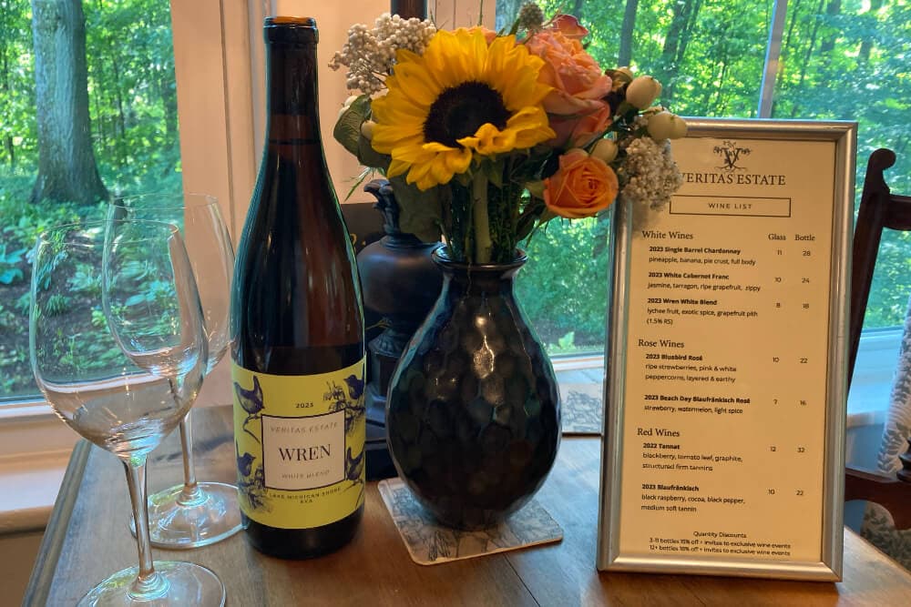A bottle of Wren wine sits next to a vase of flowers and a wine list on a table by a window.
