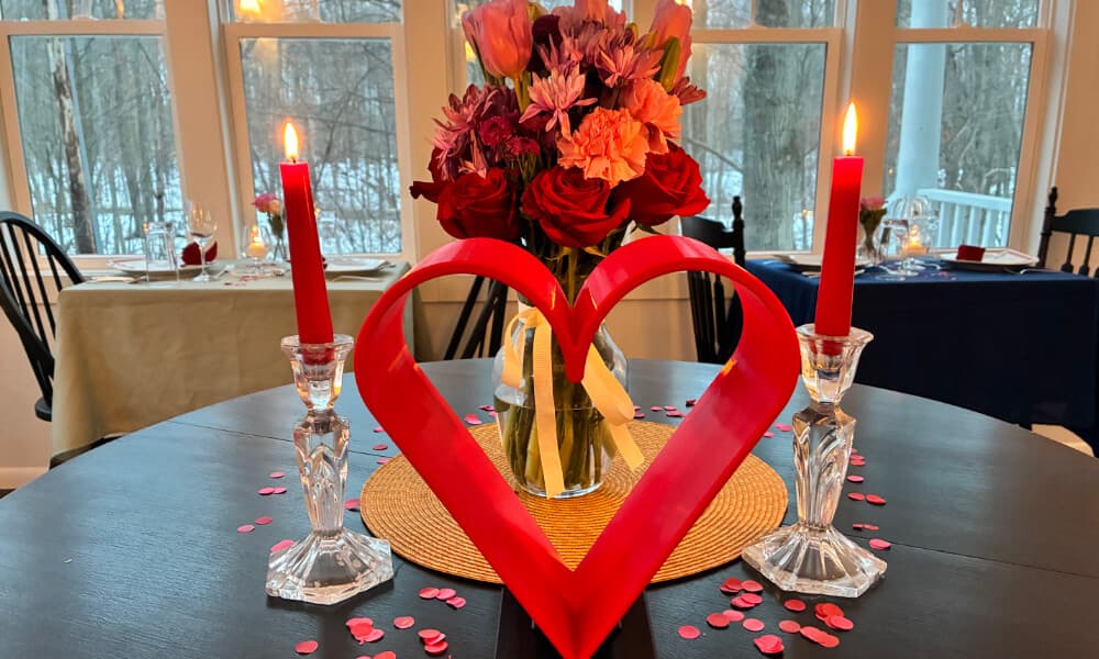 A romantic dinner table set with a heart-shaped decoration, red candles, and flowers, surrounded by scattered petals.