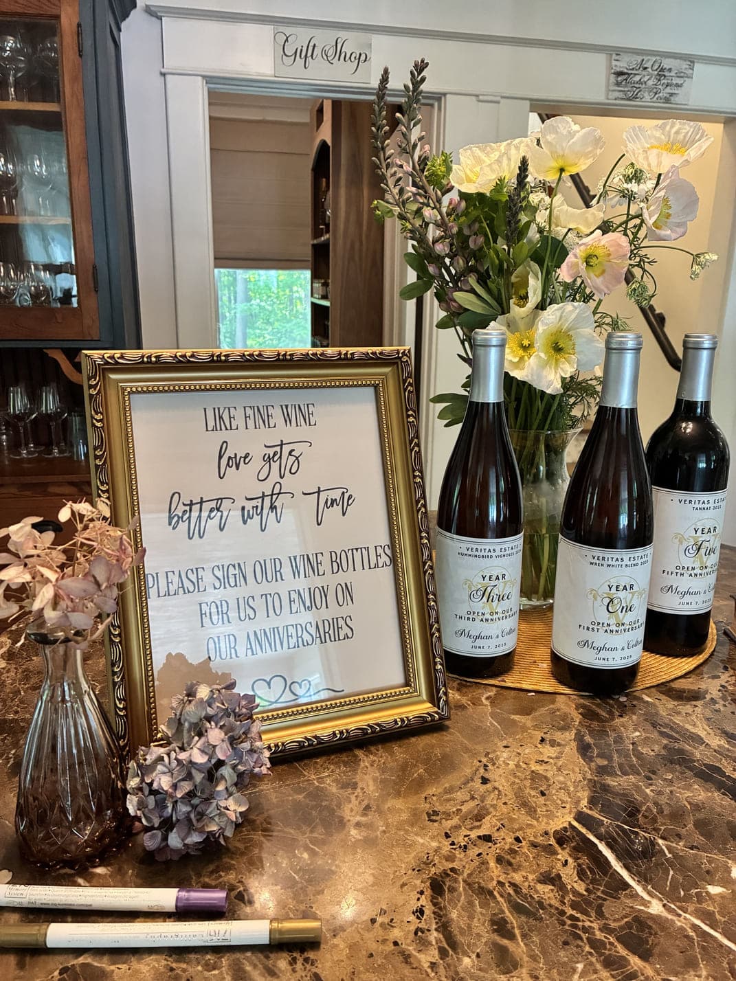 A decorative sign invites guests to sign wine bottles alongside three wine bottles and a bouquet of flowers on a table.