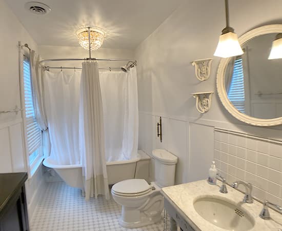 A bright, white vintage-style bathroom featuring a classic clawfoot soaking tub with a wraparound shower curtain, a marble-top vanity, and a round mirror reflecting a warm pendant light.