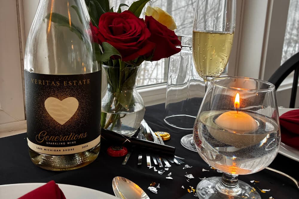 A cozy table setting featuring a bottle of Veritas Estate Generations sparkling wine, red roses, champagne flutes, and a candle.