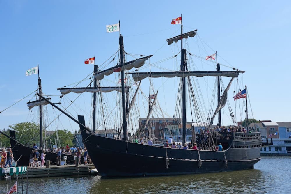 A tall ship with numerous flags and people aboard docked at a waterfront.