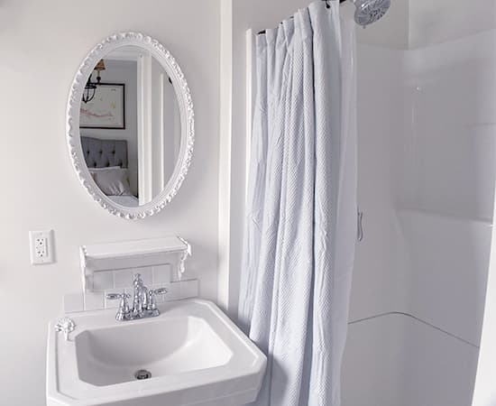 A minimalist bathroom featuring a white pedestal sink and a matching white oval-framed mirror. The walk-in shower is enclosed with a simple white curtain, maintaining a clean and bright aesthetic.