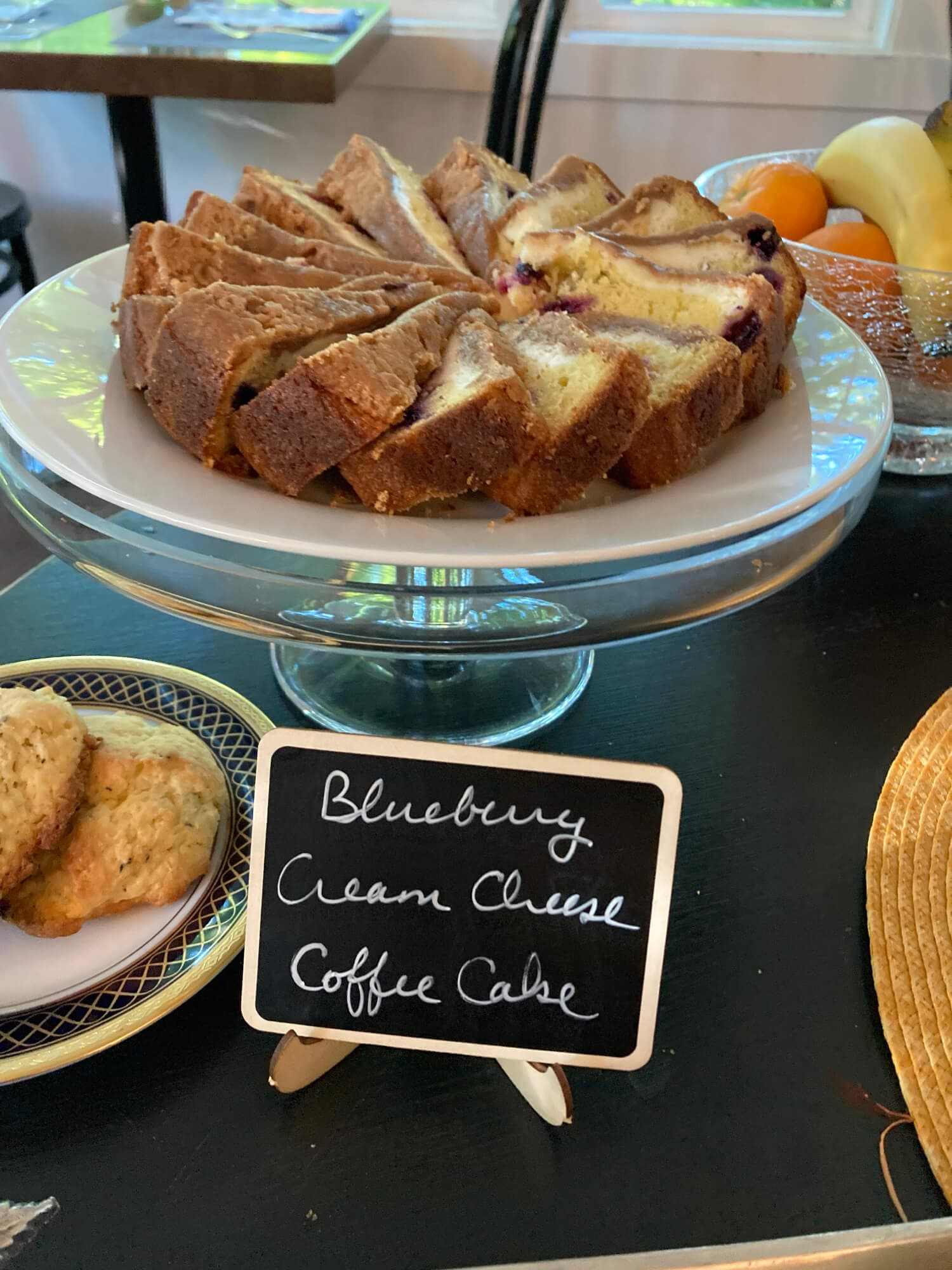 A sliced blueberry cream cheese coffee cake on a glass cake stand with a decorative sign.