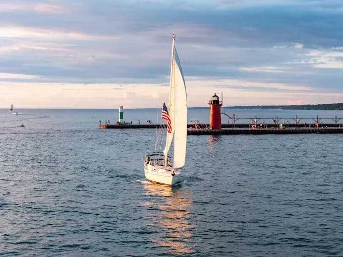 A sailboat with an American flag glides across the water near a lighthouse and pier at sunset.