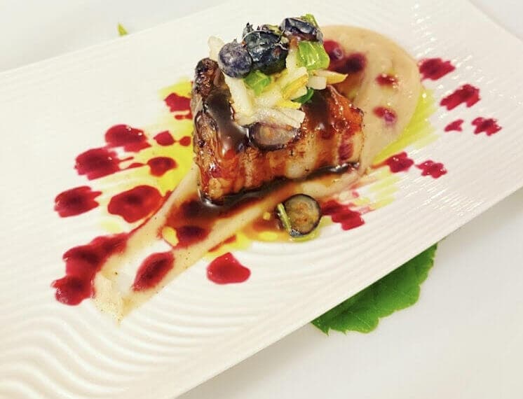 A plate with a piece of prk cooked perfectly on top of a bed of mashed potatoes, garnished with greens and vegetables, and drizzled with a bright red sauce around it